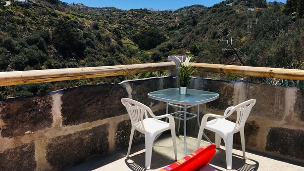 Un balcón con una mesa de vidrio y dos sillas blancas, con vistas a un paisaje montañoso.