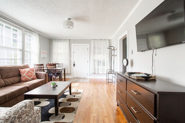 Un salon moderne avec un canapé, une table basse et un téléviseur. Il y a également un coin repas avec des chaises et une table. La pièce est dotée d’un parquet et d’une grande fenêtre.