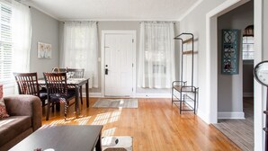 Una sala de estar con piso de madera, una mesa de comedor con sillas, un sofá y una mesa de centro. Se observa una puerta con marco blanco y una ventana con cortinas blancas.