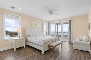 A bedroom with a bed, a nightstand, a bench, a ceiling fan, and a view of the ocean through a sliding glass door.