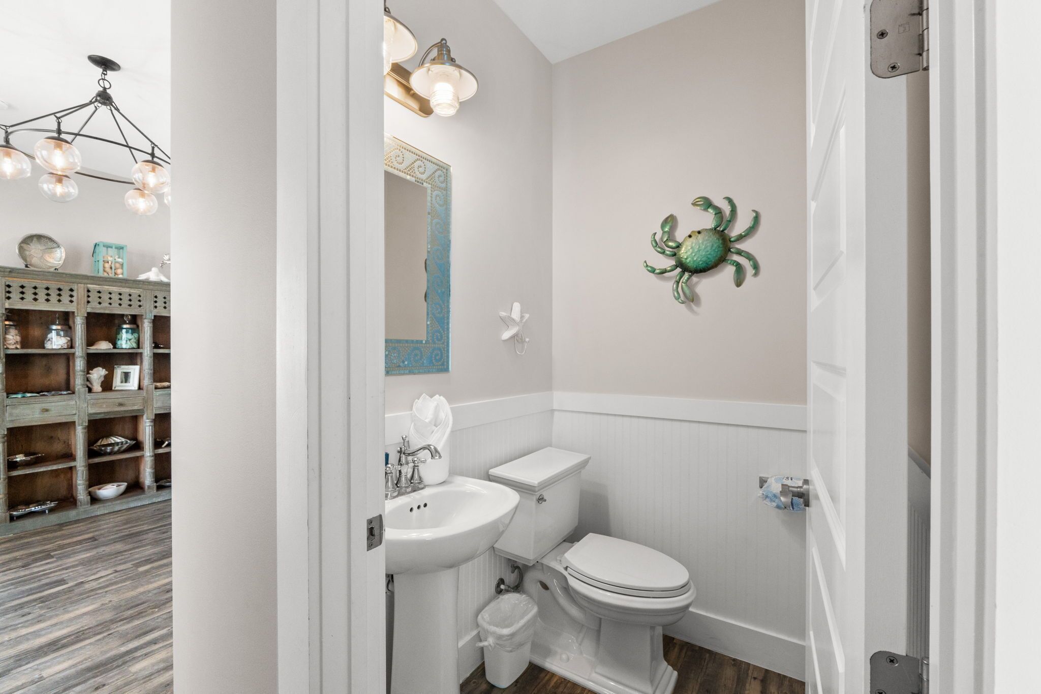 Une salle de bain avec un lavabo, des toilettes et un meuble-lavabo, tous de couleur blanche. Un crabe décoratif est accroché au mur, et un lustre se trouve au-dessus du lavabo.