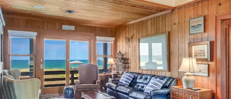 A cozy living room with a leather sofa, armchairs, and a wooden coffee table. Large windows offer a view of the ocean.