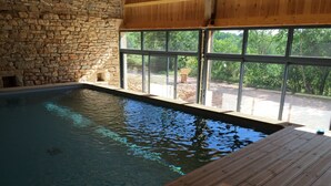 Une piscine intérieure aux murs en pierre et au sol en bois, de grandes fenêtres offrant une vue sur la verdure.