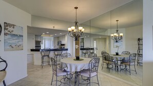 A dining area with a round table, chairs, and a chandelier. There is a kitchen in the background and a large mirror on the wall. - 503 Barrington Arms - Crisp and Beachy.  What a View! (Hilton Head Island)