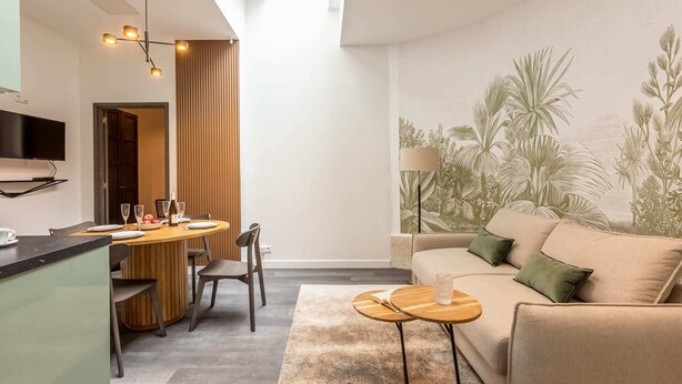 A modern living room with a sofa, a dining area with a table and chairs, a TV mounted on the wall, and a large window.