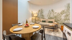 A dining area with a round wooden table, six chairs, and a sofa with a green pillow. A bottle of wine and a bowl of fruit are on the table.