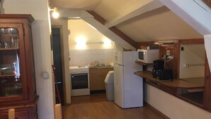 Eine kompakte Küche mit Herd, Backofen, Mikrowelle und Kühlschrank. In der Ecke befindet sich ein Holzschrank mit Geschirr und einem Besen.