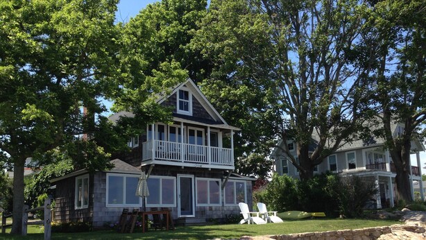 Une maison à deux étages avec un balcon, entourée d’arbres et d’un mur de pierre.
