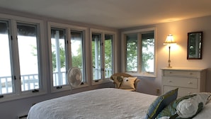 Une chambre avec un grand lit, un ventilateur sur pied, une chaise avec des coussins, une commode et une fenêtre donnant sur des arbres.