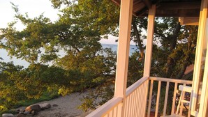 Une terrasse en bois avec une table et des chaises, donnant sur une plage bordée de rochers et d’arbres.