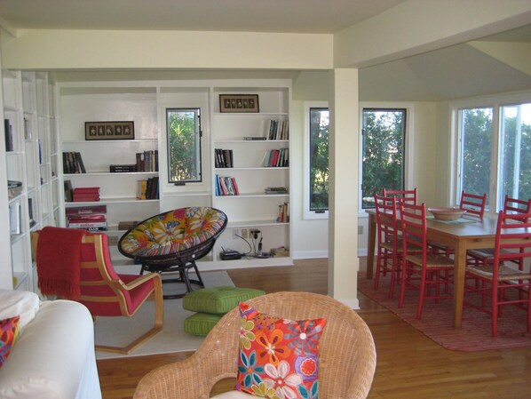 A living room with a dining area, a bookshelf, and large windows.