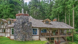 Una casa grande y moderna con chimenea de piedra, múltiples ventanas y un porche cubierto con mobiliario exterior.