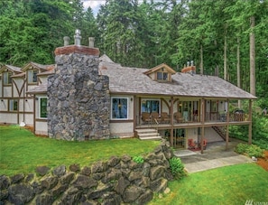 Ein großes, modernes Haus mit einem Steinkamin, zahlreichen Fenstern und einer Veranda mit Sitzgelegenheit.