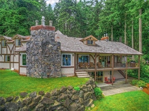 A large, modern house with a stone chimney, multiple windows, and a covered porch with outdoor seating.