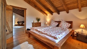Un dormitorio con techo de madera, una cama con ropa de cama floral, una mesita de noche, una lámpara y una cesta de mimbre.