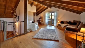 Un loft espacioso con vigas de madera, una escalera, un rincón de descanso acogedor con un sofá y un sillón, y un gran ventanal que aporta luz natural.