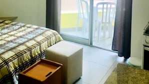 Quarto com cama, uma mesinha, um banquinho, janela com cortinas e vista para o exterior.