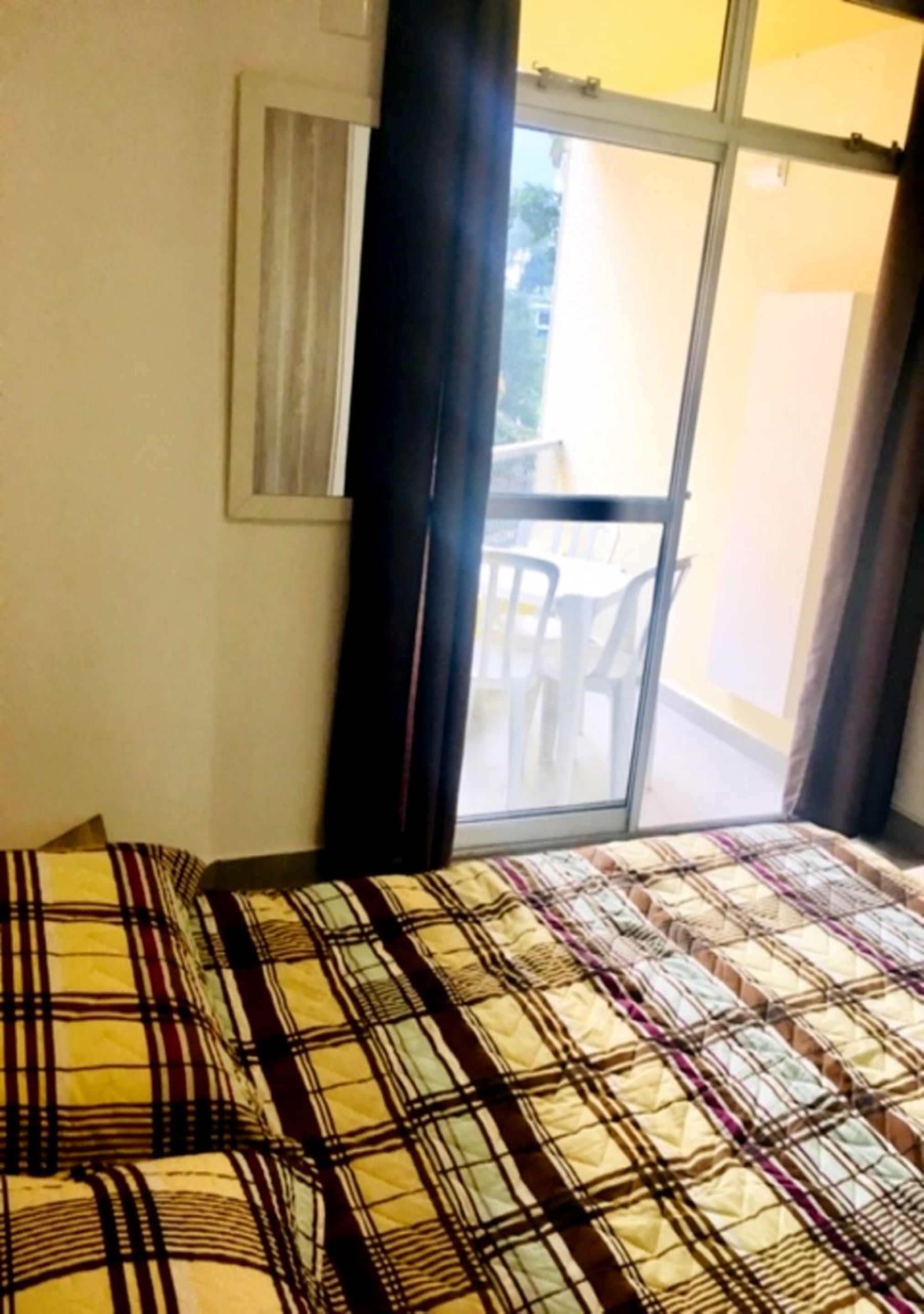 A room with a bed, a window with curtains, and a view of an outdoor area with chairs.