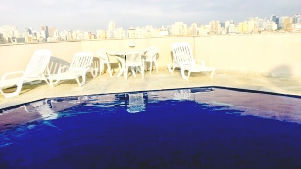 Piscina no terraço com cadeiras de sol brancas e vista para o horizonte da cidade.