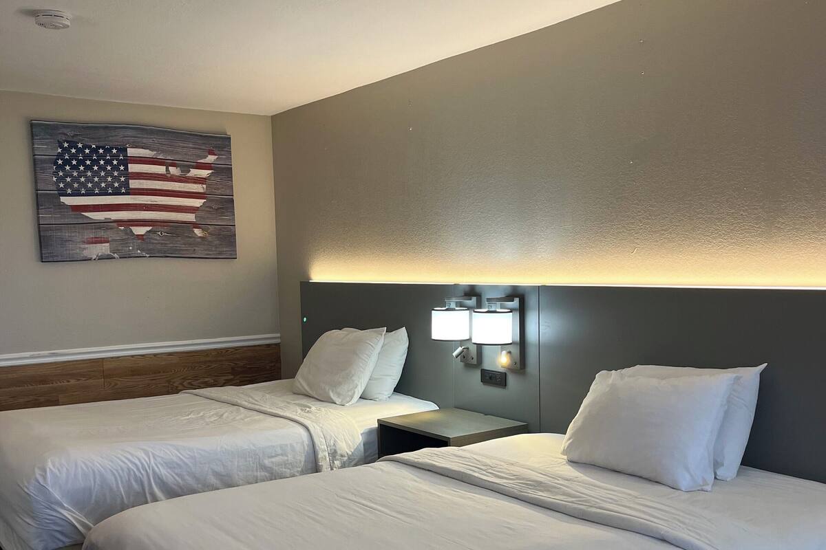 A hotel room with two beds, a nightstand with lamps, and a wall-mounted American flag artwork.