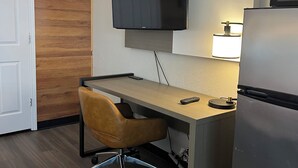 Habitación de hotel compacta con pared de acento de madera, un televisor de pantalla plana sobre un escritorio, un refrigerador pequeño y un cesto de basura.