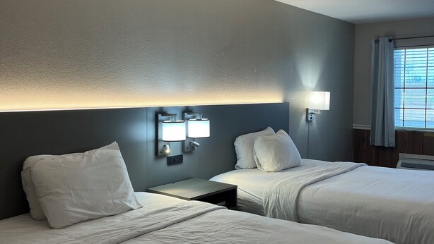Habitación de hotel con dos camas, una mesita de noche, lámparas de pared y un ventanal con cortinas.
