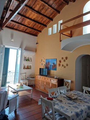 A room with a wooden ceiling, a television on a wooden cabinet, white chairs, a table with a patterned tablecloth, and a view of the sea through an open door.