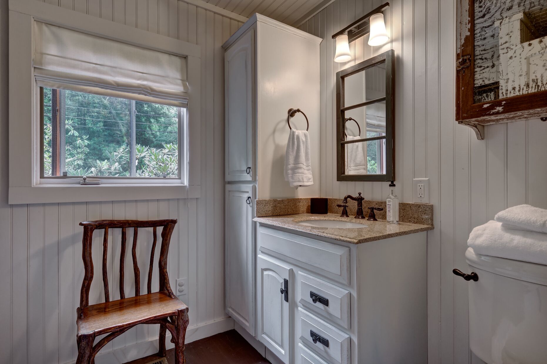 Um banheiro com uma cadeira de madeira, pia com espelho e janela com vista para o verde.