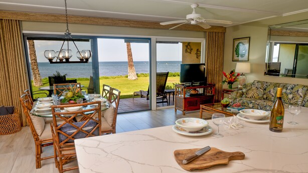 A spacious living area with a large island table, dining set, and a sofa. A television is mounted on the wall, and there is a view of the ocean through the glass doors.