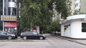 Une vue de rue avec un bâtiment portant l’inscription « LUNEDOR » et un autre bâtiment avec une enseigne « FARMOP ». On aperçoit deux voitures garées et un arbre au premier plan.