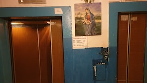 Un ascenseur avec des portes en bois et un mur bleu orné d’une peinture religieuse et d’une liste de numéros d’urgence.