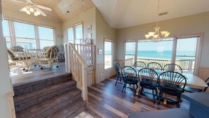 A spacious room with a wooden staircase, a dining area with chairs and a table, and large windows offering a view of the ocean. - Finding Sandity | 5BR 4x4 OBX | Pool, Hot Tub & Game Rm | Sleeps 10 (Carova)