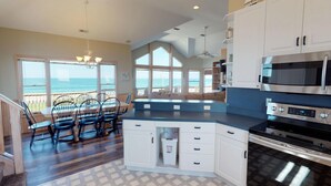 A kitchen with a large island, white cabinets, and stainless steel appliances. There is a dining area with chairs and a view of the ocean through large windows. - Finding Sandity | 5BR 4x4 OBX | Pool, Hot Tub & Game Rm | Sleeps 10 (Carova)