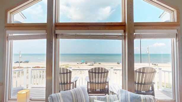 A room with large windows overlooking a beach, featuring two striped chairs and a view of the ocean. - Finding Sandity | 5BR 4x4 OBX | Pool, Hot Tub & Game Rm | Sleeps 10 (Carova)