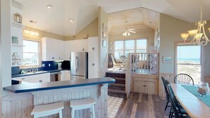 A modern kitchen with white cabinets, stainless steel appliances, and a blue countertop. There is a dining area with a table and chairs, and a view of the beach through large windows. - Finding Sandity | 5BR 4x4 OBX | Pool, Hot Tub & Game Rm | Sleeps 10 (Carova)