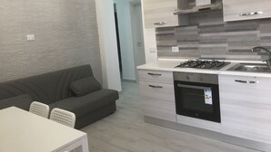 A modern kitchen with a gas stove, sink, and dishwasher. There is a grey sofa and a white table with chairs in the living area.