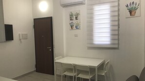 A small, well-lit room with a white table and four chairs, a door, an air conditioning unit, and a window with blinds.