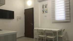 A small, modern kitchen with a white countertop, a dark brown door, and a white dining table with two chairs. There is an air conditioning unit above the door and a wall-mounted TV on the left.