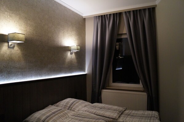 A hotel room with a headboard, two wall-mounted lights, a window with curtains, and a radiator.