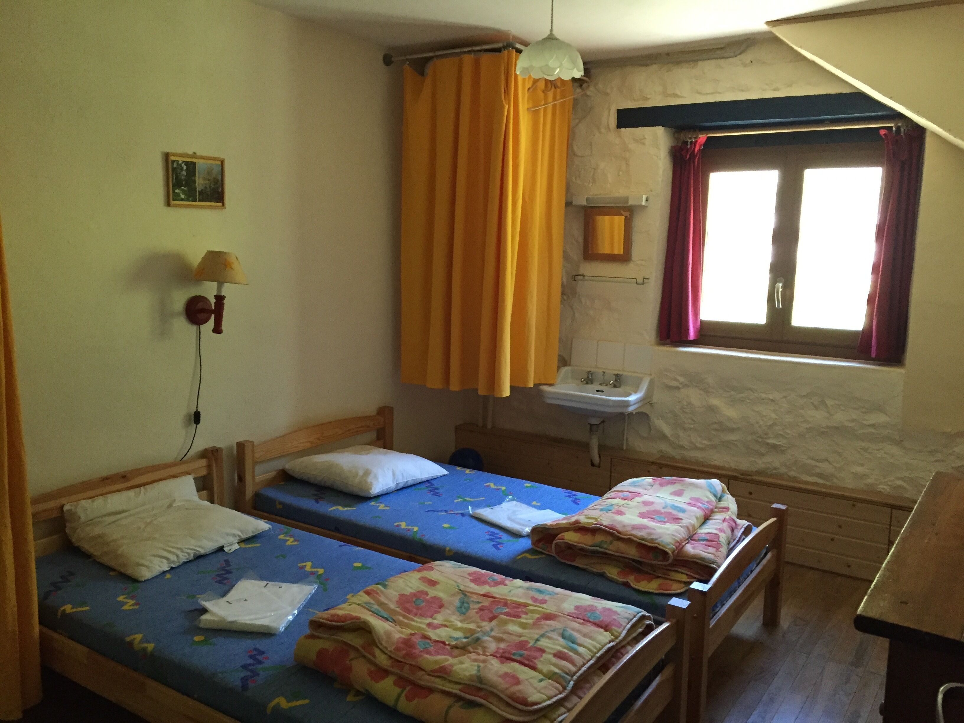 A small, simple room with two single beds, a sink, and a window with curtains.