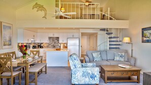 A spacious living area with a dining table, a sofa, and a coffee table. A kitchen is visible in the background with white cabinets and a refrigerator. - Maalaea Surf, Unit A5-2 Bedroom Beachfront Townhouse, AC, Wifi, TV, Kitchen (Kihei)