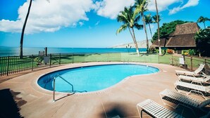 A swimming pool with a curved edge, surrounded by a fence, and overlooking a beach with palm trees and a clear sky. - Maalaea Surf, Unit A5-2 Bedroom Beachfront Townhouse, AC, Wifi, TV, Kitchen (Kihei)