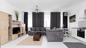 Una sala de estar moderna con un sofá gris, mueble de entretenimiento de madera y una zona de cocina con armarios blancos y una estufa.