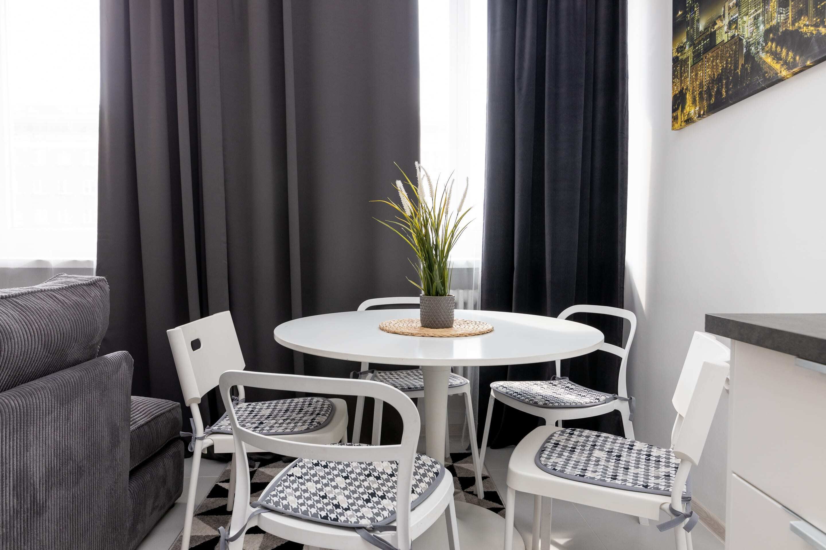 Un salon moderne avec une table ronde blanche, quatre chaises blanches dotées de coussins à motifs, un canapé gris et une plante en pot posée sur la table.