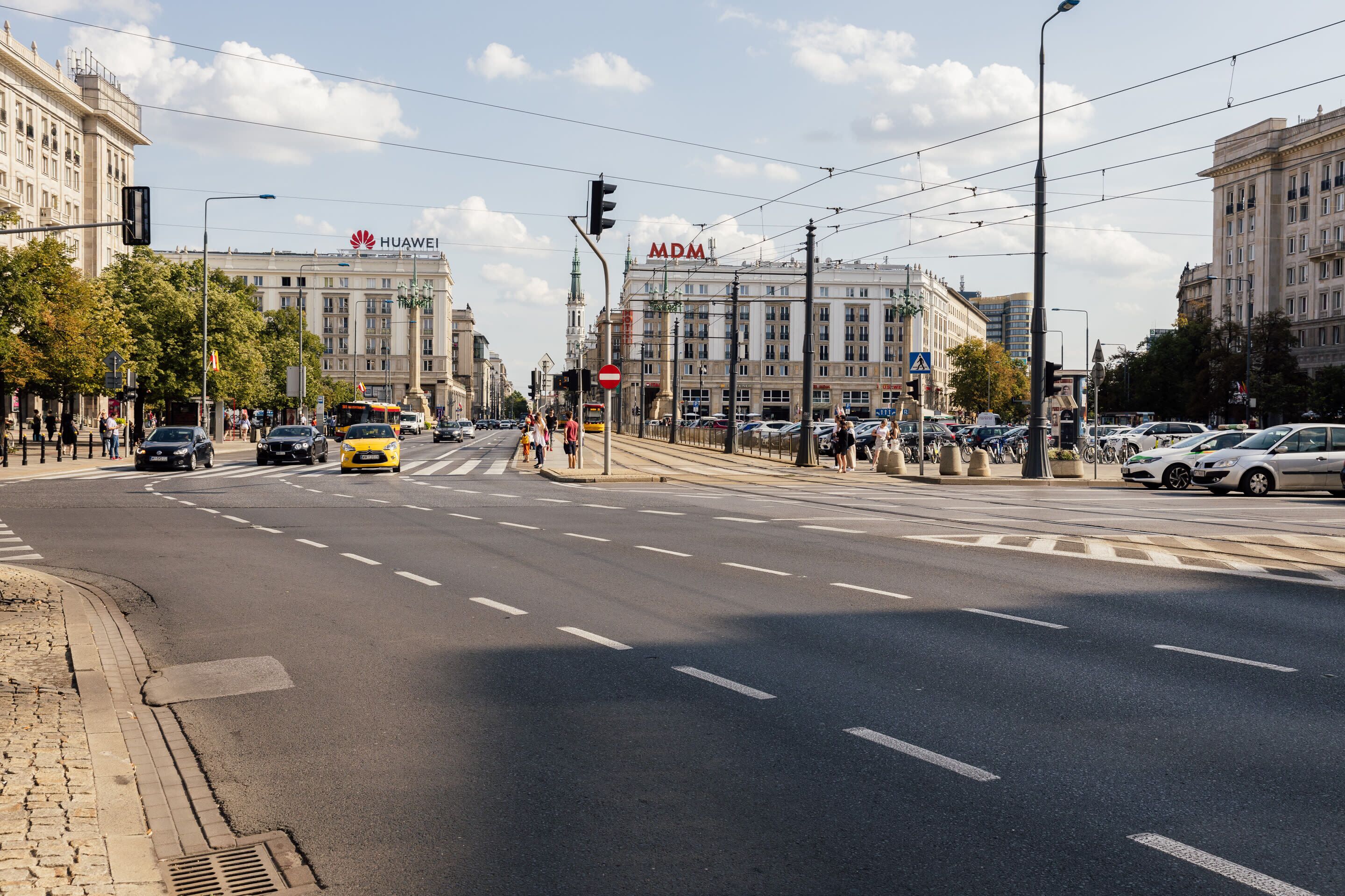 A wide urban street with buildings, including a prominent Huawei sign, and a tram.