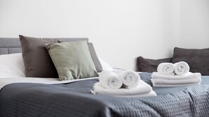 Una cama bien hecha con ropa de cama y almohadas grises, y una pila de toallas enrolladas sobre la cama.