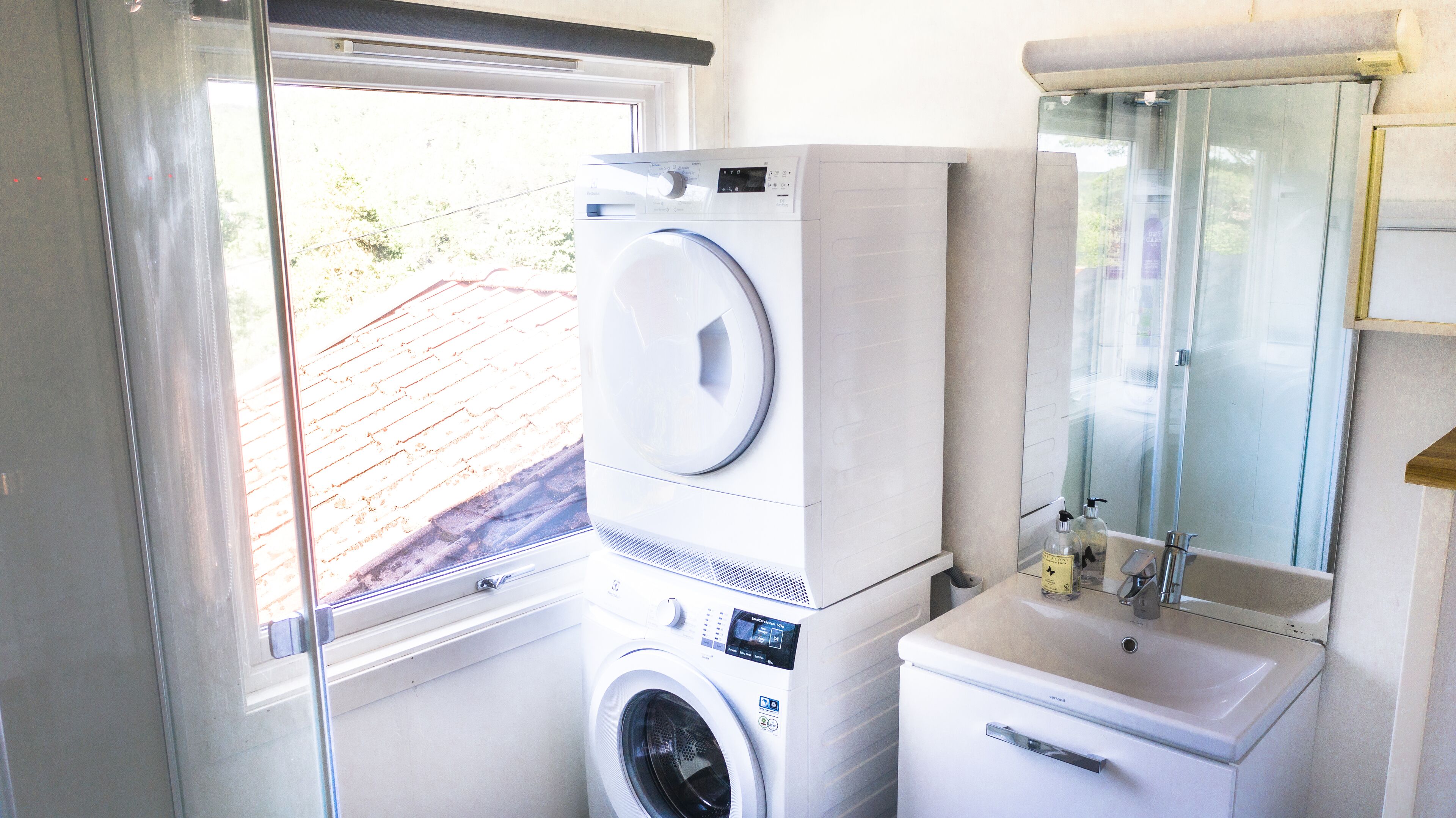 A compact laundry area with a front-loading washer and dryer, a sink with a mirror above it, and a window providing natural light.