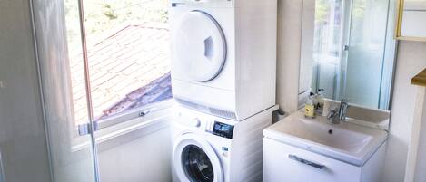 A compact laundry area with a front-loading washer and dryer, a sink with a mirror above it, and a window providing natural light.