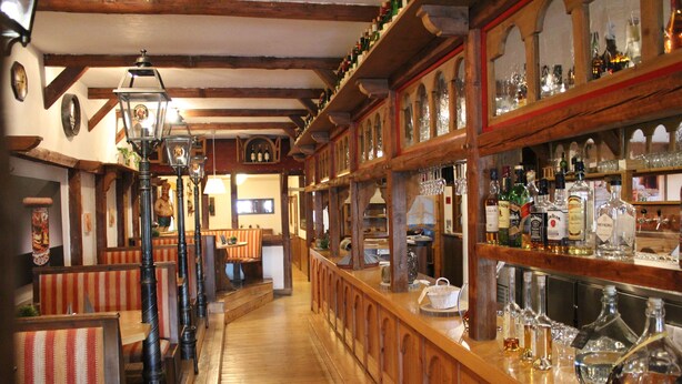 A bar area with wooden beams, a long counter with bottles and glasses, and striped benches. - Hotel - Restaurant Franziskaner am Markt (Loningen)