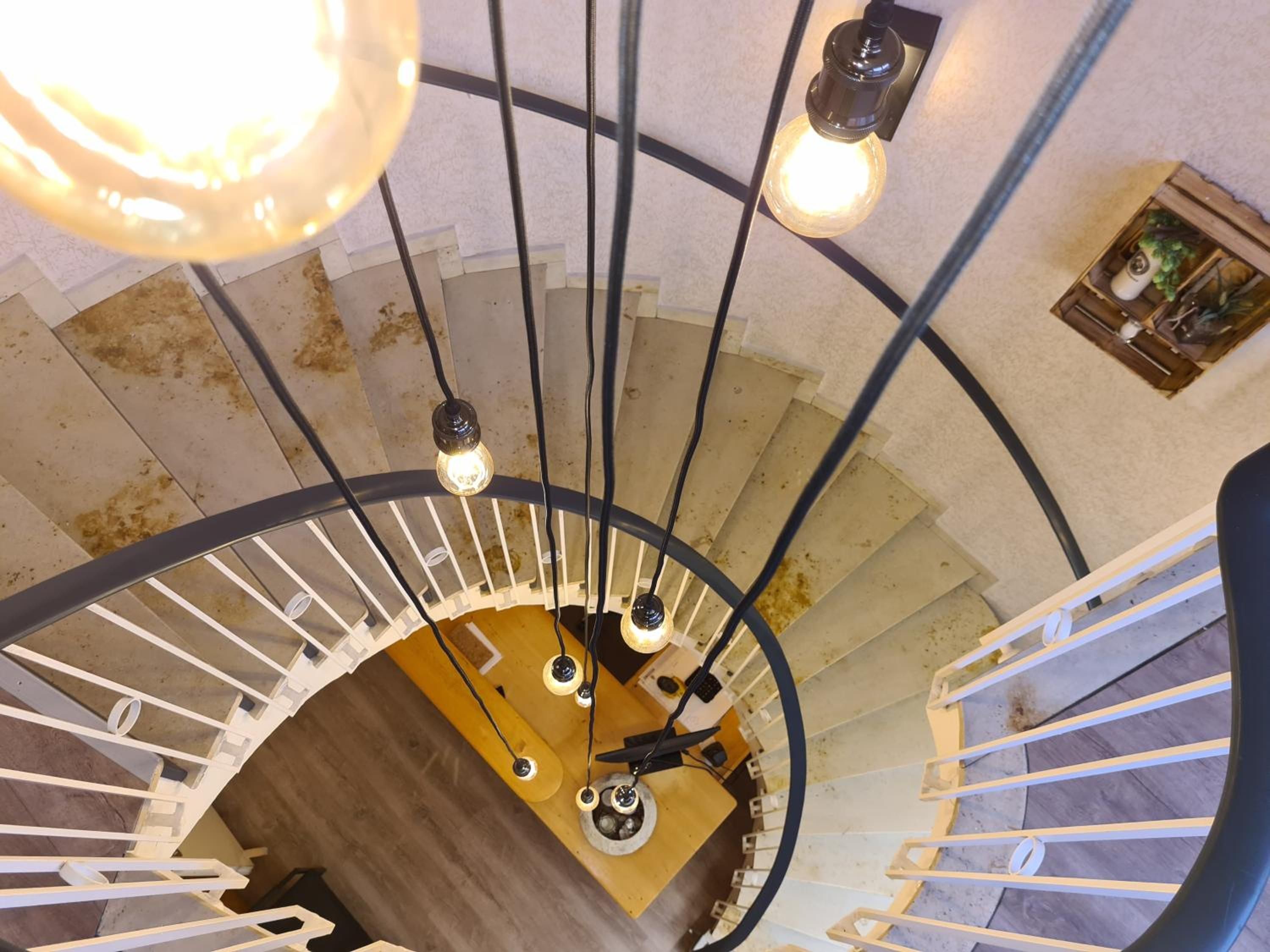 Una escalera de caracol con una plataforma central de madera, barandillas de metal negro y lámparas colgantes.